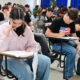 Estudantes realizam prova em sala de aula, sentados individualmente em carteiras enquanto respondem às questões impressas.
