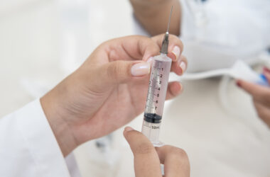 Mãos seguram uma seringa com agulha durante atividade prática na área da saúde, com uma pessoa de jaleco ao fundo.