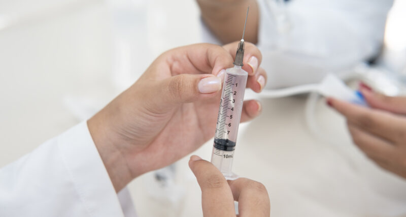 Mãos seguram uma seringa com agulha durante atividade prática na área da saúde, com uma pessoa de jaleco ao fundo.