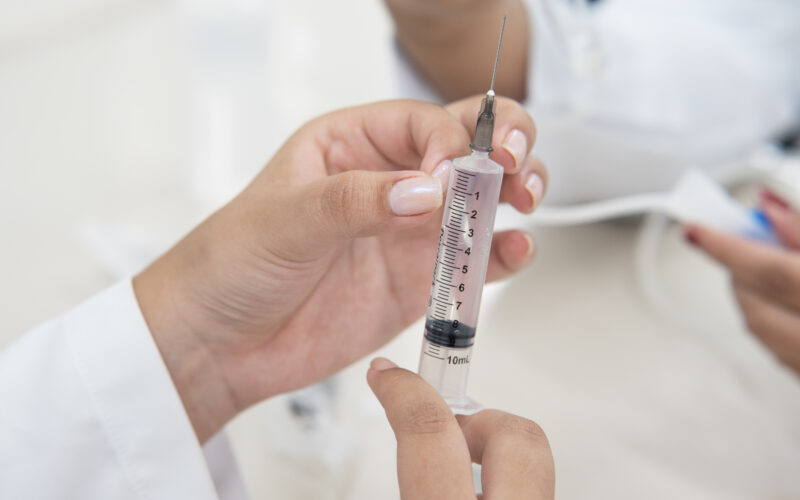 Mãos seguram uma seringa com agulha durante atividade prática na área da saúde, com uma pessoa de jaleco ao fundo.