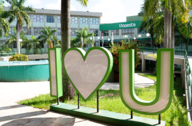 Campus da Unoeste em Presidente Prudente, com letreiro verde e branco “I love U” em primeiro plano, cercado por palmeiras, jardins e área de convivência. Ao fundo, aparece o prédio da universidade com a identificação Unoeste na fachada.