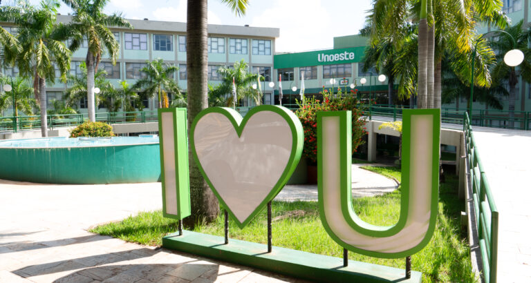 Campus da Unoeste em Presidente Prudente, com letreiro verde e branco “I love U” em primeiro plano, cercado por palmeiras, jardins e área de convivência. Ao fundo, aparece o prédio da universidade com a identificação Unoeste na fachada.