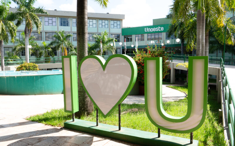 Campus da Unoeste em Presidente Prudente, com letreiro verde e branco “I love U” em primeiro plano, cercado por palmeiras, jardins e área de convivência. Ao fundo, aparece o prédio da universidade com a identificação Unoeste na fachada.
