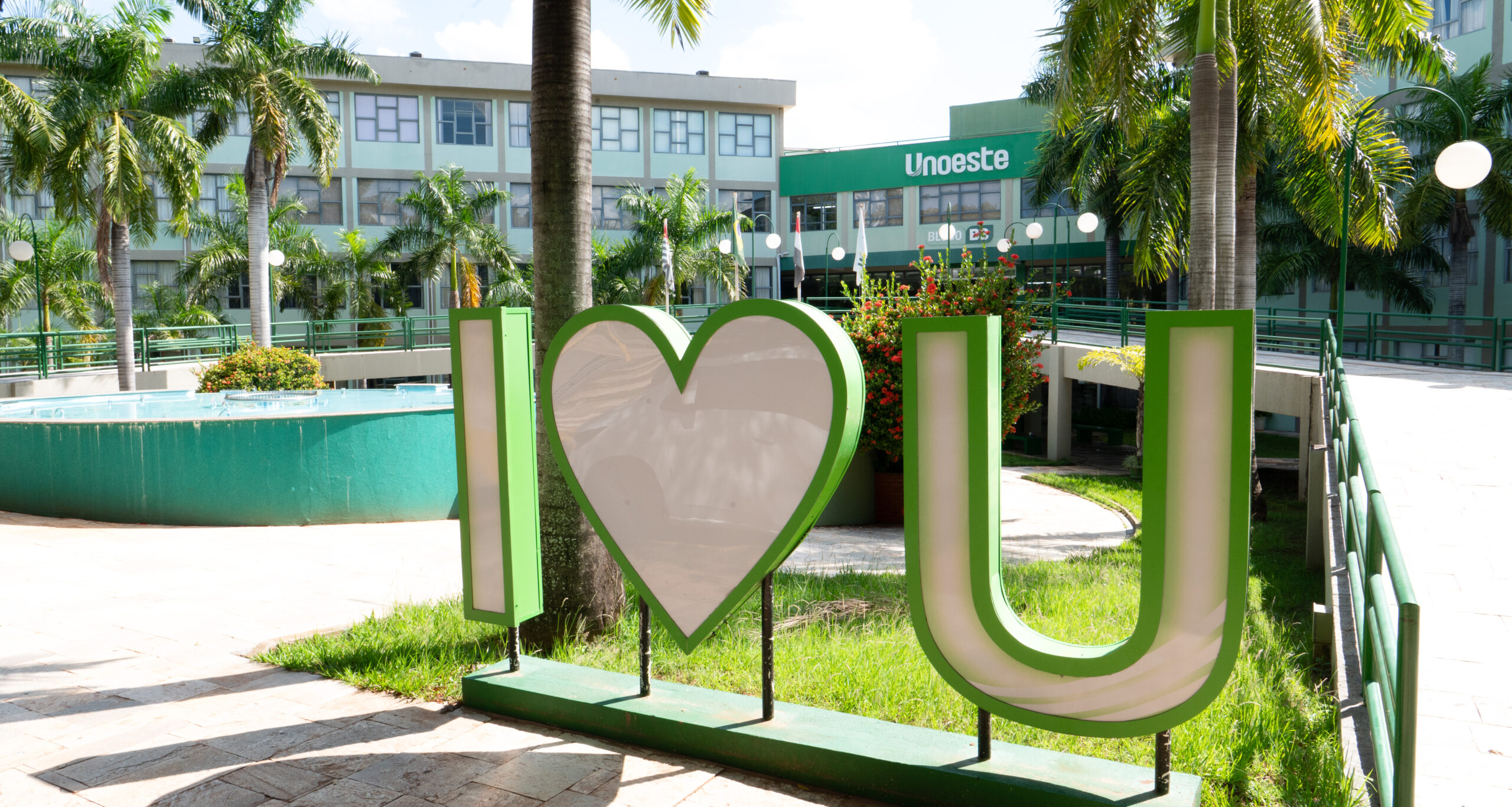 Campus da Unoeste em Presidente Prudente, com letreiro verde e branco “I love U” em primeiro plano, cercado por palmeiras, jardins e área de convivência. Ao fundo, aparece o prédio da universidade com a identificação Unoeste na fachada.