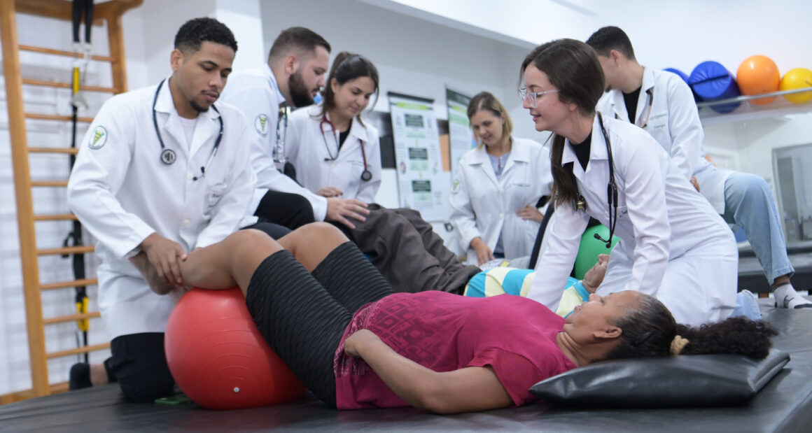 Estudantes de Fisioterapia realizam atendimento prático com pacientes em laboratório, utilizando bola terapêutica durante exercícios de reabilitação.