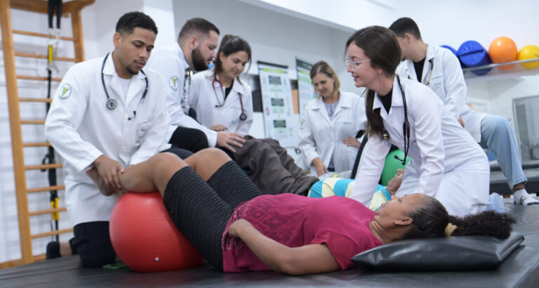 Estudantes de Fisioterapia realizam atendimento prático com pacientes em laboratório, utilizando bola terapêutica durante exercícios de reabilitação.