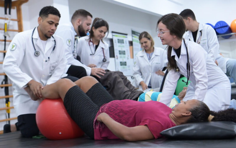 Estudantes de Fisioterapia realizam atendimento prático com pacientes em laboratório, utilizando bola terapêutica durante exercícios de reabilitação.