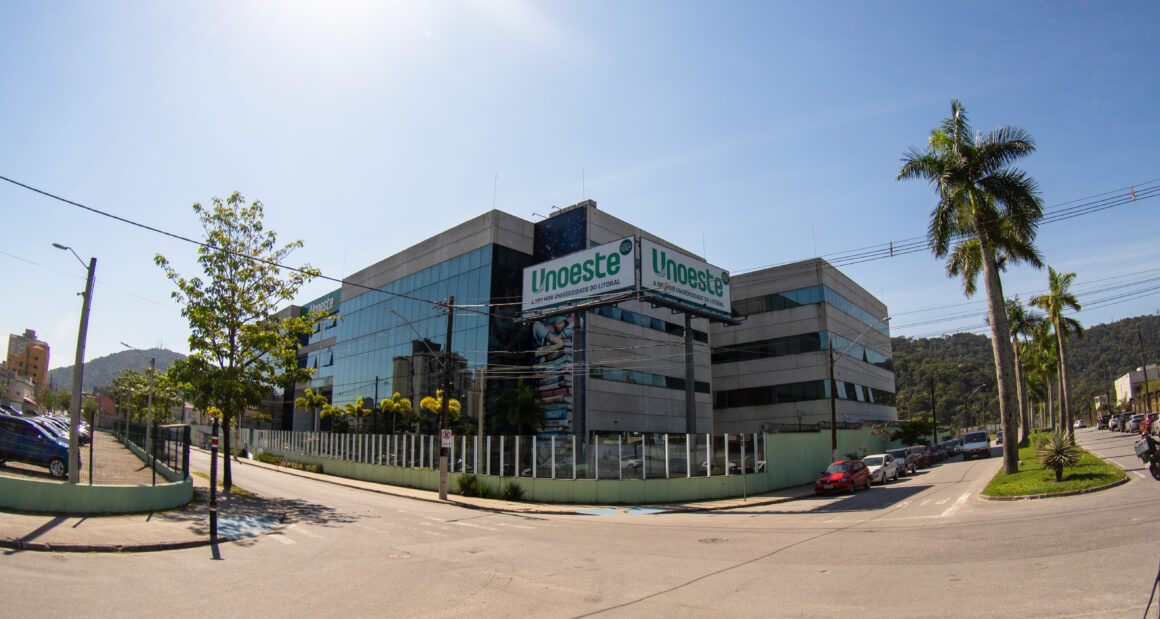 Vista externa do campus da Unoeste em Guarujá, com fachada de vidro e concreto, placas com o nome da universidade e palmeiras ao redor.