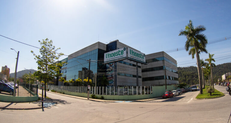 Vista externa do campus da Unoeste em Guarujá, com fachada de vidro e concreto, placas com o nome da universidade e palmeiras ao redor.