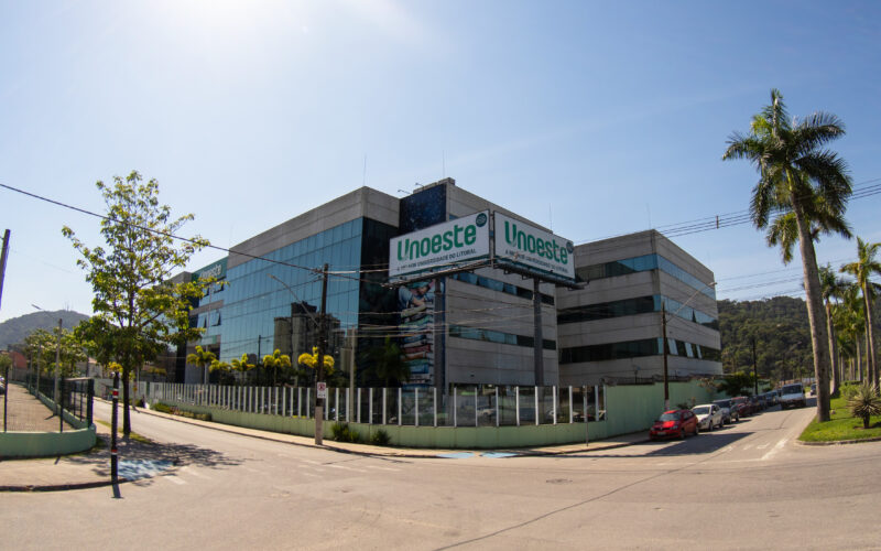 Vista externa do campus da Unoeste em Guarujá, com fachada de vidro e concreto, placas com o nome da universidade e palmeiras ao redor.