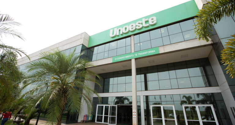 Fachada do campus da Unoeste em Jaú, com estrutura moderna de vidro, letreiro da universidade em destaque, entrada principal e palmeiras ao redor do prédio.