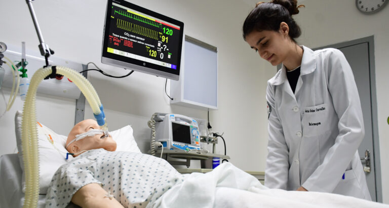 Estudante de Enfermagem da Unoeste realiza atividade prática em laboratório de simulação, ao lado de manequim hospitalar conectado a equipamentos de monitoramento.