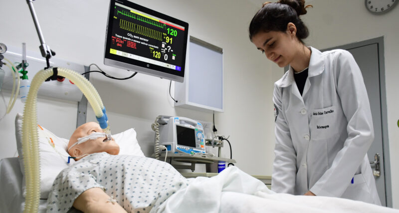Estudante de Enfermagem da Unoeste realiza atividade prática em laboratório de simulação, ao lado de manequim hospitalar conectado a equipamentos de monitoramento.