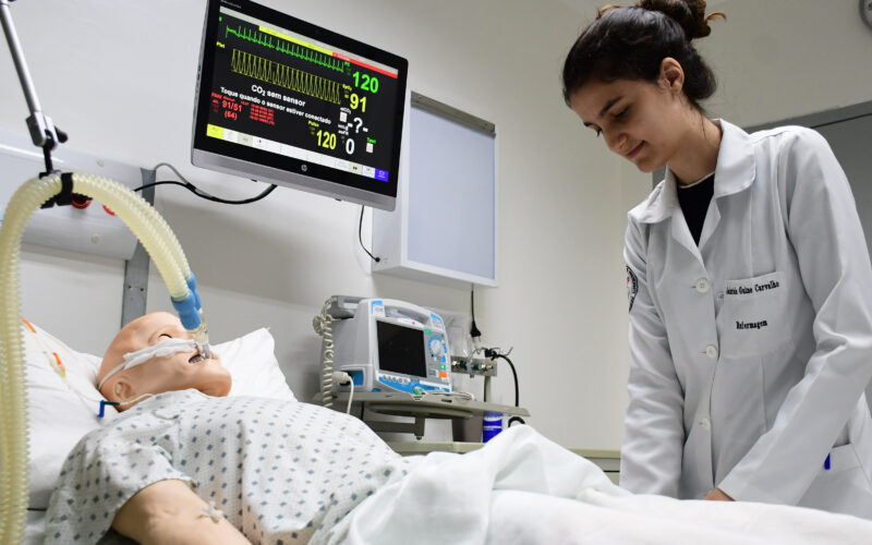 Estudante de Enfermagem da Unoeste realiza atividade prática em laboratório de simulação, ao lado de manequim hospitalar conectado a equipamentos de monitoramento.