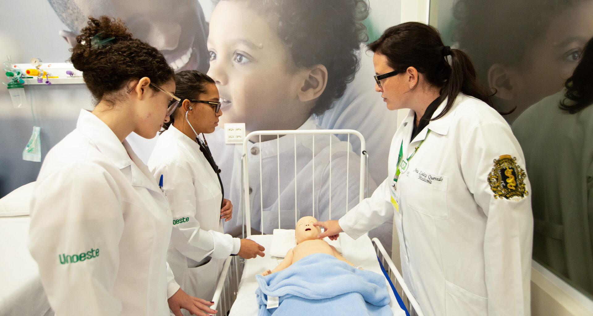 Estudantes de Enfermagem da Unoeste participam de aula prática com professora, realizando simulação de cuidados com um manequim de bebê em laboratório.