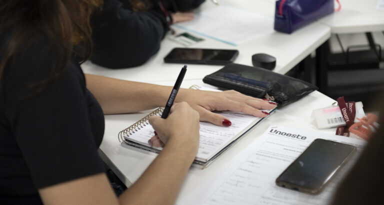Estudante faz anotações em caderno durante atividade em sala de aula, com materiais da Unoeste sobre a mesa.