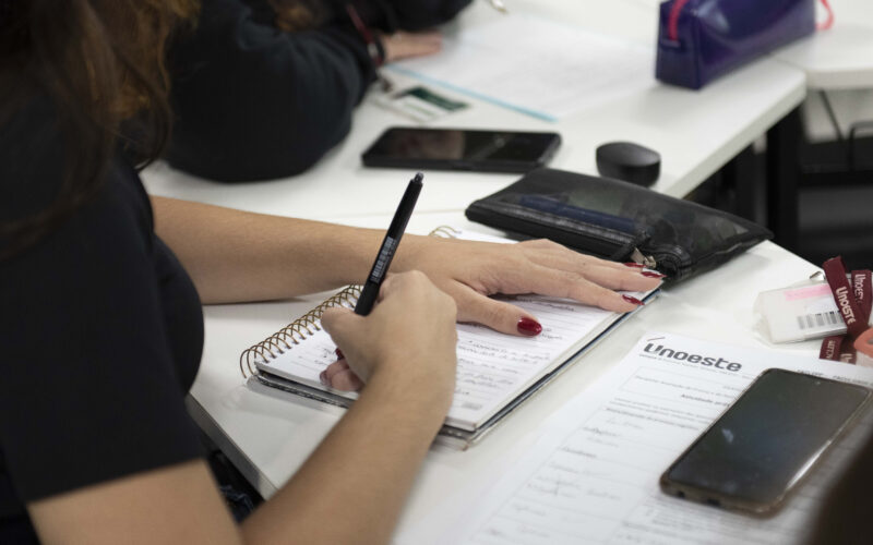 Estudante faz anotações em caderno durante atividade em sala de aula, com materiais da Unoeste sobre a mesa.