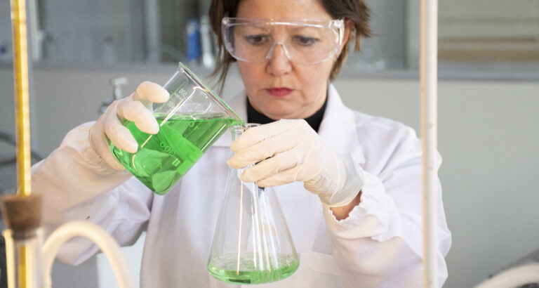 Profissional de jaleco, luvas e óculos de proteção realiza experimento em laboratório da Unoeste, transferindo uma solução verde de um béquer para um frasco de vidro.