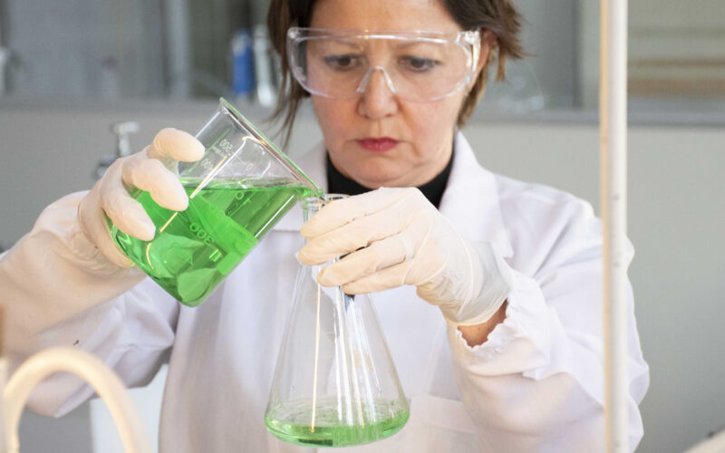 Profissional de jaleco, luvas e óculos de proteção realiza experimento em laboratório da Unoeste, transferindo uma solução verde de um béquer para um frasco de vidro.