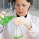 Profissional de jaleco, luvas e óculos de proteção realiza experimento em laboratório da Unoeste, transferindo uma solução verde de um béquer para um frasco de vidro.
