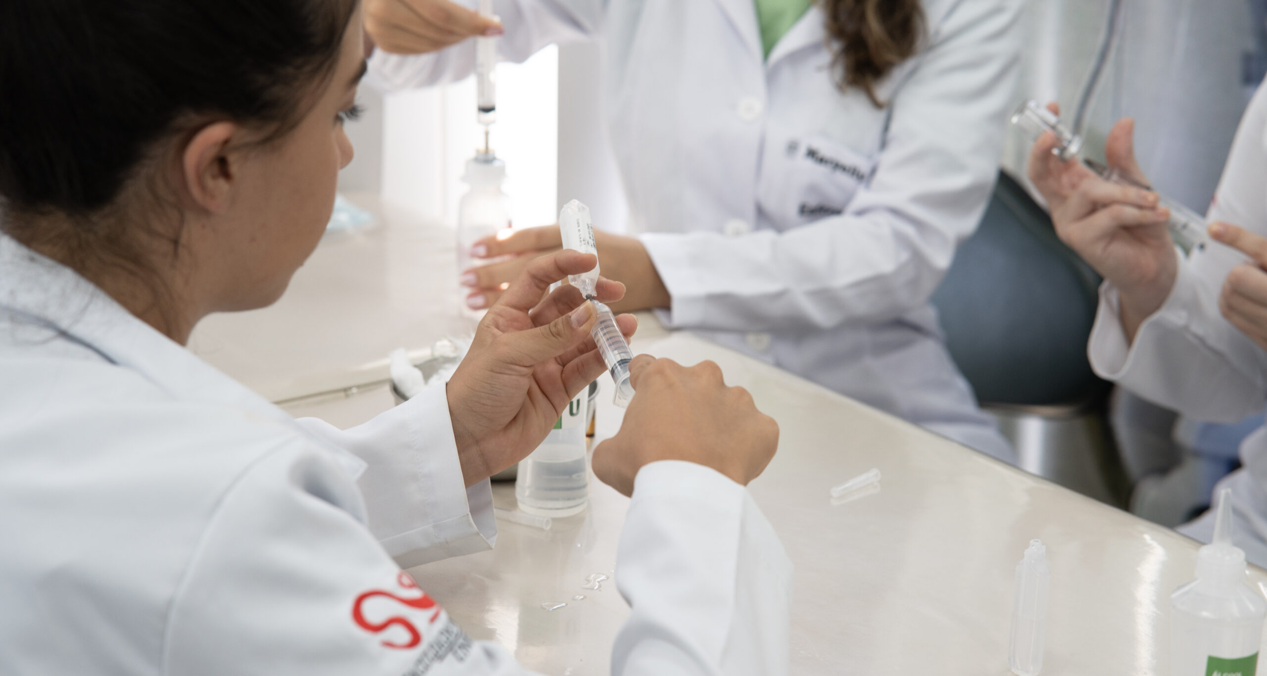 Estudantes de Enfermagem da Unoeste, usando jalecos brancos, praticam o manuseio de seringas durante atividade em laboratório.